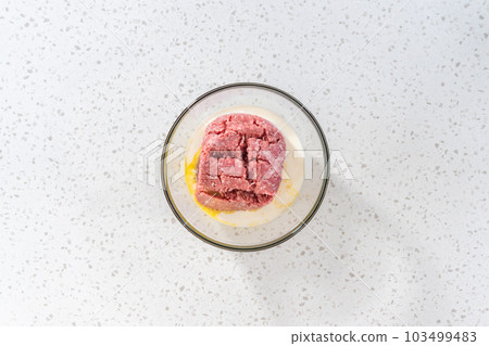 Flat lay. Mixing ingredients with a potatoe masher in a glass mixing bowl to prepare oven-baked meatballs. 103499483