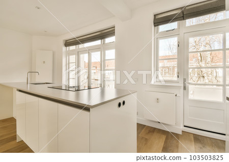 a kitchen area with white cabinets and wood flooring in an apartment, showing the large windows that look out onto the street 103503825
