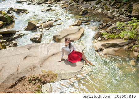 Girl in a blouse and skirt sitting near a mountain river 103507819