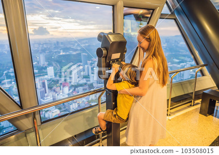 Mom and son are looking at Kuala lumpur cityscape. Use binoculars. Panoramic view of Kuala Lumpur city skyline evening at sunset skyscrapers building in Malaysia. Traveling with kids concept Mom and son are looking at Kuala lumpur cityscape. Use binoculars. Panoramic view of Kuala Lumpur city skyline evening at sunset skyscrapers building in Malaysia. Traveling with kids concept 103508015