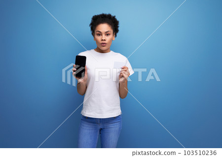 a beautiful young swarthy woman with fluffy curly hair dressed in a white t-shirt holds a smartphone a beautiful young swarthy woman with fluffy curly hair dressed in a white t-shirt holds a smartphone 103510236