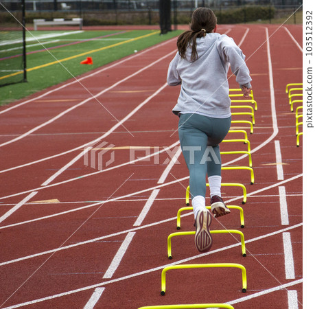 Rear view of female runner wearing spandex running over six inch mini hurdles on a track 103512392