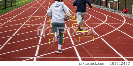 Two runners running over six iinch hurdles in lane on a track Two runners running over six iinch hurdles in lane on a track 103512427