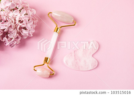 Modern selfcare concept. Pink jade roller and gua sha for face massage with flowers lilacs. Facial massager tools. Anti age, lifting and toning care. Modern selfcare concept. Pink jade roller and gua sha for face massage with flowers lilacs. Facial massager tools. Anti age, lifting and toning care. 103515016