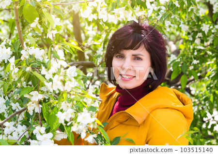 An adult brunette in a bright yellow jacket near a blooming white apple tree 103515140