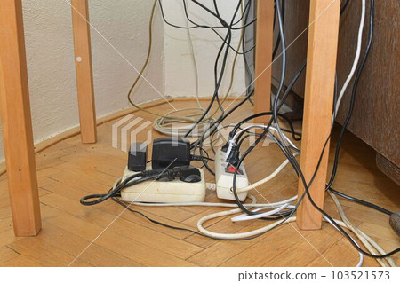 Cable chaos clutter from multiple electric wire extension cords and multi-contact plugs on wooden floor. Tangled cables on the parquet floor. 103521573