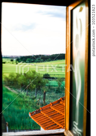Vertical photo of an open window in a rustic house overlooking green meadows with fresh grass on a spring summer day. Authentic landscape. View from the window on the street, nature. Rural scene. Vertical photo of an open window in a rustic house overlooking green meadows with fresh grass on a spring summer day. Authentic landscape. View from the window on the street, nature. Rural scene. 103521623