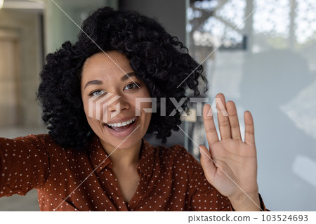 Beautiful business woman with curly hair wearing looking at smartphone camera, waving her hand in greeting gesture, talking with friends and colleagues, using video call app Beautiful business woman with curly hair wearing looking at smartphone camera, waving her hand in greeting gesture, talking with friends and colleagues, using video call app 103524693