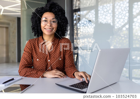 Young beautiful african american woman working inside modern office, businesswoman smiling and looking at camera at work using laptop. 103524703