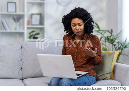Upset and worried African American young woman talking with her psychologist, psychotherapist online via laptop. Sits at home on the couch and consults via video call. 103524790