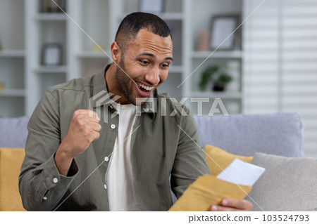 Happy hispanic young male student is happy to receive news. Sits at home on the couch and reads letters, documents, exam results, tests. Shows a victory gesture with his hand. 103524793
