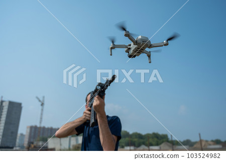 A man aims to shoot a rifle at a flying drone against a blue sky. A man aims to shoot a rifle at a flying drone against a blue sky. 103524982