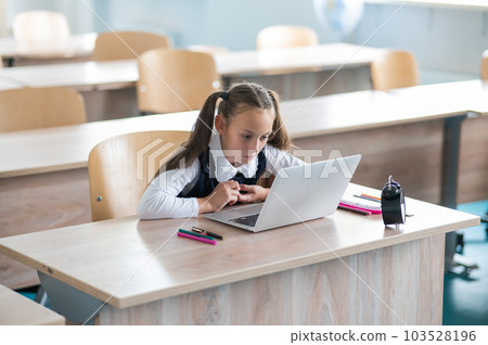 Caucasian girl sits at a desk at school and studies at a laptop. Alarm clock on the table. 103528196