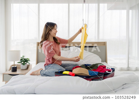 Young asian woman checking casual tank top t-shirt before putting it in suitcase. A luggage full of clothes and hat for summer vacation trip are wide open on a bed. Young asian woman checking casual tank top t-shirt before putting it in suitcase. A luggage full of clothes and hat for summer vacation trip are wide open on a bed. 103528249