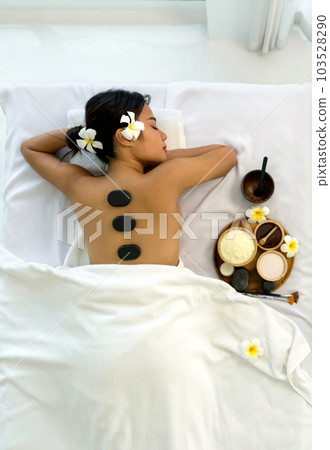 Woman getting spa hot stones massage. Lying on a massage table, relaxing with eyes closed. Spa Thai therapy treatment aromatherapy for beauty body. Spa beauty massage healthy wellness. Top View Woman getting spa hot stones massage. Lying on a massage table, relaxing with eyes closed. Spa Thai therapy treatment aromatherapy for beauty body. Spa beauty massage healthy wellness. Top View 103528290