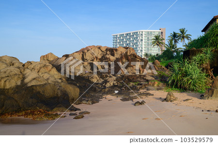 Tropical beach picturesque landscape photograph, rocky sandy beach, and the urban hotel building in the background. Clear blue skies. Tropical beach picturesque landscape photograph, rocky sandy beach, and the urban hotel building in the background. Clear blue skies. 103529579