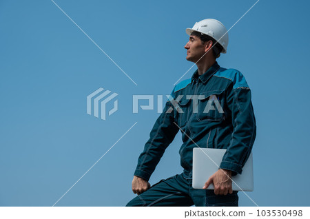 A male builder in a helmet stands against a blue sky with a laptop in his hands.  103530498