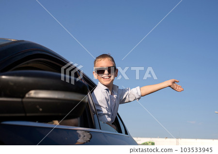 A cute boy in sunglasses looked out of the car window waving his hand with a laugh A cute boy in sunglasses looked out of the car window waving his hand with a laugh 103533945