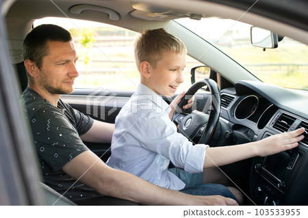 Dad and happy son in the front seat of a car driving a car. The boy holds the steering wheel. 103533955