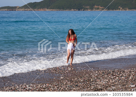 Young brunette woman with long hair in a white pareo and hat walks along the beach. 103535984