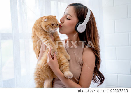 Pretty Asian girl wear earphone and hold and kiss cat in front of glass windows with white curtain Pretty Asian girl wear earphone and hold and kiss cat in front of glass windows with white curtain 103537213
