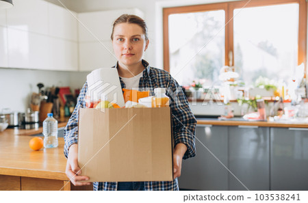 Young redhead woman carrying a delivery box full of fresh organic groceries in the kitchen. A donation box of different products on house background. Express food delivery, online shopping concept 103538241
