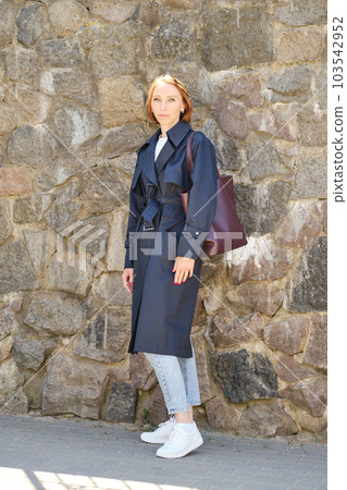 Full length portrait of redhead woman wearing trench coat, jeans and sneakers 103542952