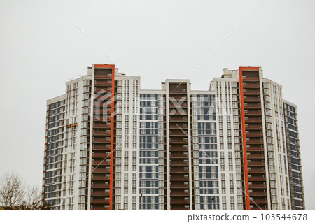A white and red facade of a modern multi-story residential building on a construction site. New construction in a big city, megalopolis. Tall house with lots of windows front view low angle photo. 103544678