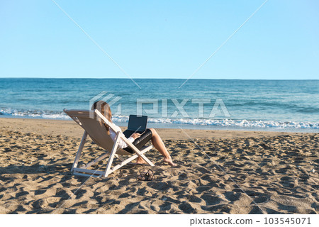 Back view of alone young brunette woman freelancer in office clothes with a laptop looking on the sea sitting on the deck chair on beach. Summer concept Back view of alone young brunette woman freelancer in office clothes with a laptop looking on the sea sitting on the deck chair on beach. Summer concept 103545071