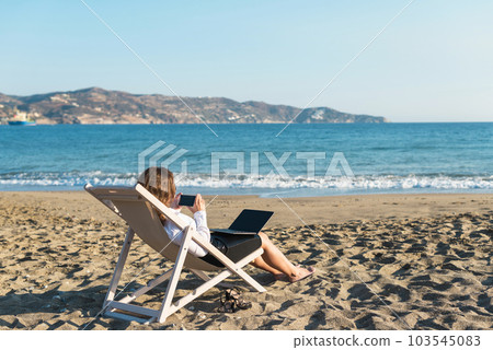 Back view of alone young brunette woman freelancer in office clothes with a laptop looking on the sea sitting on the deck chair on beach. Summer concept 103545083