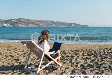 Back view of alone young brunette woman freelancer in office clothes with a laptop looking on the sea sitting on the deck chair on beach. Summer concept Back view of alone young brunette woman freelancer in office clothes with a laptop looking on the sea sitting on the deck chair on beach. Summer concept 103545084