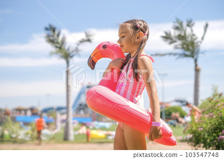 A girl in a pink swimsuit holds a swimming circle in the shape of a flamingo looking away. Summer vacation at the water park. A girl in a pink swimsuit holds a swimming circle in the shape of a flamingo looking away. Summer vacation at the water park. 103547748