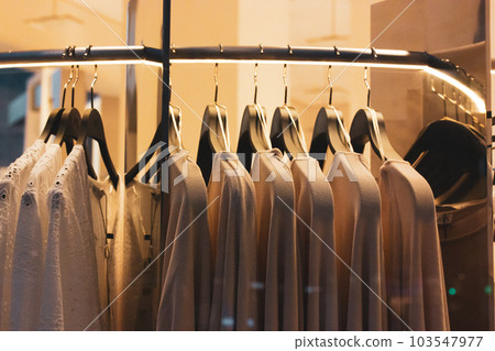 Women's clothing store. White blouses hang on hangers in a store window with artificial warm light. Beauty, fashion business concept. Female clothes shirt, sweater, sundress, dresses. Apparel rack. 103547977