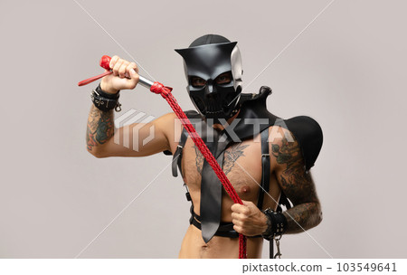 sexy a man in a bdsm skull mask, dressed in a leather raincoat with leather handcuffs and straps on the body on a light background of a copy paste 103549641
