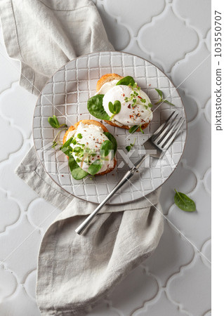 Poached egg sandwich with cream cheese, spinach and micro greens. Healty breakfast bruschetta on linen napkin white tile background. Keto diet, text space, top view Poached egg sandwich with cream cheese, spinach and micro greens. Healty breakfast bruschetta on linen napkin white tile background. Keto diet, text space, top view 103550707