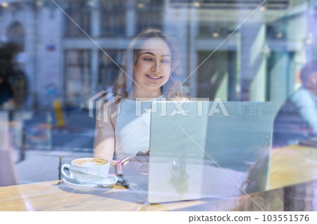Beautiful girl inside cafe with cup of coffee by laptop looking through window Beautiful girl inside cafe with cup of coffee by laptop looking through window 103551576