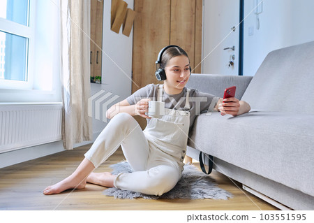 Young female teenager in headphones sitting at home on floor near sofa looking in smartphone 103551595