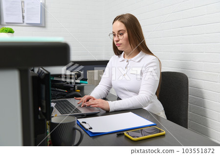 Confident female receptionist, office manager working on laptop, sitting behind a reception desk in modern clinic, keeping appointments with medical staff. Healthcare worker at outpatient hospital Confident female receptionist, office manager working on laptop, sitting behind a reception desk in modern clinic, keeping appointments with medical staff. Healthcare worker at outpatient hospital 103551872