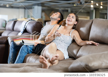 In store wife settled down with legs to test of sofa in use,husband examines upholstery fabric In store wife settled down with legs to test of sofa in use,husband examines upholstery fabric 103556325