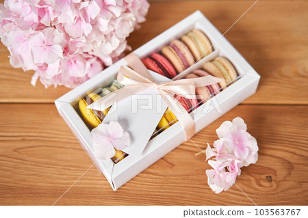 Dessert French Macarons in present box. Sweet dessert on a flowers background. Still life with macaroni cakes and different flower. Delivery concept, coffee and flower shop Dessert French Macarons in present box. Sweet dessert on a flowers background. Still life with macaroni cakes and different flower. Delivery concept, coffee and flower shop 103563767