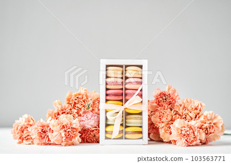 Dessert French Macarons in present box. Sweet dessert on a flowers background. Still life with macaroni cakes and different flower. Delivery concept, coffee and flower shop Dessert French Macarons in present box. Sweet dessert on a flowers background. Still life with macaroni cakes and different flower. Delivery concept, coffee and flower shop 103563771