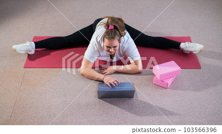 Young caucasian fat woman doing bends on a sports mat and watching a training video on a laptop. A chubby girl stretches the split remotely using video communication Young caucasian fat woman doing bends on a sports mat and watching a training video on a laptop. A chubby girl stretches the split remotely using video communication 103566396