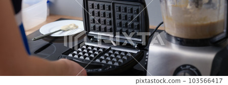 Woman opens waffle iron to make waffles in kitchen. Woman opens waffle iron to make waffles in kitchen. 103566417