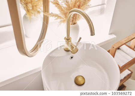 A beautiful sink with a golden faucet next to an oval mirror and a shelf with hand towels. Close-up of an elegant golden faucet in the bathroom sink next to stylish decorations. 103569272