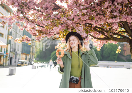 Attractive young female tourist photographer with stylish clothes and a boho hat holds bagel obwarzanek traditional polish cuisine snack walking around on blossoming sakura trees background in Krakow 103570640
