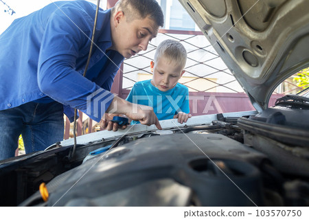 Dad shows his son where to fill in the coolant in the engine compartment. Topping up antifreeze in the car cooling system. Expansion tank near the engine. Red liquid 103570750
