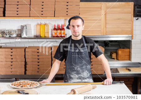 The chef looks at the camera. Raw pizza ready to bake. Cook in a blue apron in the kitchen. with a shovel in his hands. boxes for food delivery on background. The chef looks at the camera. Raw pizza ready to bake. Cook in a blue apron in the kitchen. with a shovel in his hands. boxes for food delivery on background. 103570981