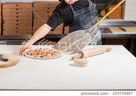Raw pizza ready to bake. Cook in a blue apron in the kitchen. with a shovel in his hands. boxes for food delivery on background. Raw pizza ready to bake. Cook in a blue apron in the kitchen. with a shovel in his hands. boxes for food delivery on background. 103570987