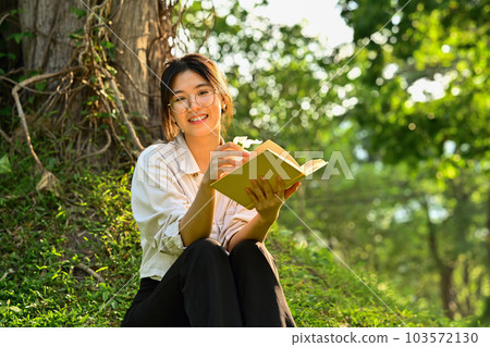 Pleased asian woman reading book under tree on green grass at sunny beautiful day Pleased asian woman reading book under tree on green grass at sunny beautiful day 103572130