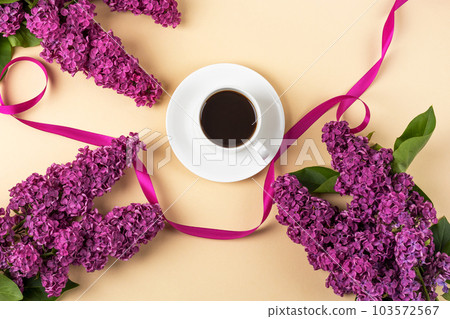 Cup of fresh black coffee and branch of blooming purple lilac with satin ribbon on orange background. Coffee break. Spring flowers. Flat lay, top view, copy space. 103572567